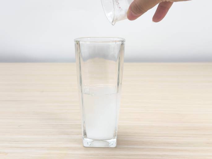 A hand pours a white powder from a small glass measuring beaker into a clear glass containing water. The glass sits on a light woodgrain table against a plain white background. The powder is dissolving into the water, creating a cloudy white mixture at the bottom of the glass. This image likely depicts a step in a science experiment or a similar activity where a powder is dissolved in water.