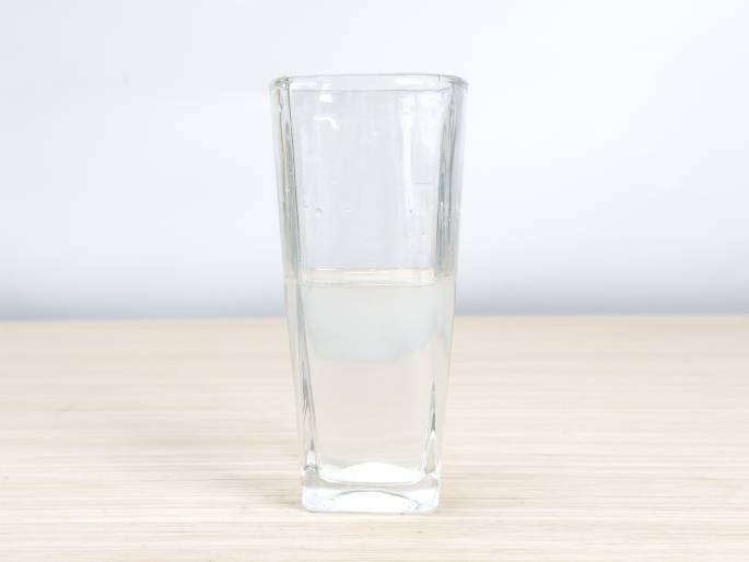 A clear glass sits on a light woodgrain surface against a plain white background. The glass is partially filled with a cloudy, white liquid, suggesting a substance has been dissolved in the water. This image likely depicts a stage in an experiment or recipe where an ingredient has been mixed into water.