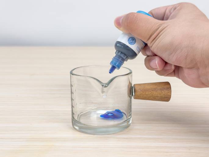 A hand holds a small bottle of blue food coloring over a clear glass measuring cup with a wooden handle. A drop of blue coloring is falling from the bottle into the cup, where a small puddle of blue liquid has already collected at the bottom. The cup sits on a light woodgrain table against a plain white background. This image likely depicts adding color to a mixture during a science experiment or cooking activity.
