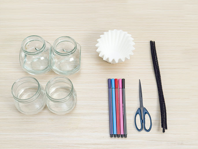 An assortment of craft supplies is arranged on a light wood table. These include four small, clear glass jars (two pairs placed side-by-side), a white coffee filter, a group of colorful markers in shades of purple, pink, red, and blue, a pair of scissors, and a black pipe cleaner. The items appear to be laid out in preparation for a craft project.