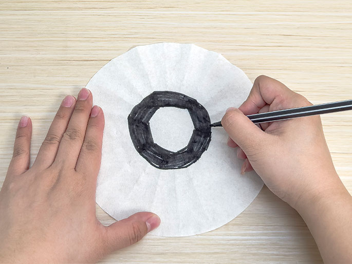 On a light wooden table, a left hand holds a white coffee filter flat while a right hand draws a thick black ring with a black marker near the center of the filter. The pleated edges of the filter are still visible.