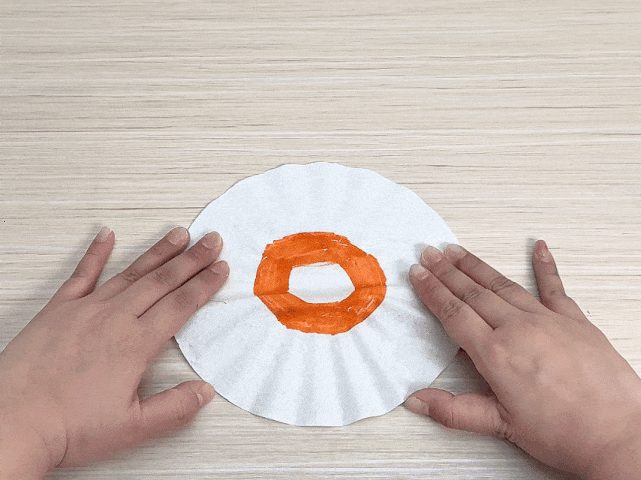 Two hands manipulate a round white coffee filter on a light woodgrain surface. An orange ring, reminiscent of a donut, has been drawn on the filter. The hands carefully fold the filter in half, then in half again, creating a triangular shape. The video demonstrates the initial steps of a craft project, likely involving the coffee filter as a base material for a decorative item.