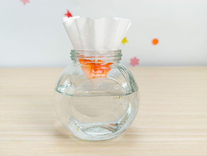 A clear glass jar filled with water sits on a light wood table. A white coffee filter rests in the jar's opening, acting as a funnel. Orange marker ink is concentrated on the filter where it dips into the water, demonstrating a chromatography experiment in progress. The background features a scattering of small, colorful star and dot stickers.