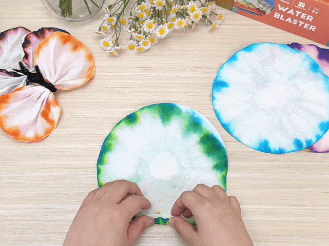 On a light wooden table, several round coffee filters are displayed. Some filters, dyed in orange and purple hues, have been crafted into a butterfly shape using a black pipe cleaner body. Other filters exhibit vibrant tie-dye patterns in blue and green. A pair of hands folds a green and blue tie-dyed filter in half, then in half again, forming a triangular shape. A small vase of daisies and a box labeled Water Blaster are also visible on the table. The video showcases a craft activity using tie-dyed coffee filters, likely to create decorative items like butterflies.