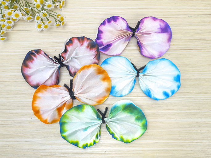 Several colorful coffee filter butterflies are arranged on a light wood surface. Each butterfly is made from two round filters, folded and tied together in the center with a black pipe cleaner to create wings. The filters are decorated with vibrant tie-dye patterns in various colors, including orange, pink and dark purple, blue, and green. A small bunch of daisies is positioned in the top left corner of the image.