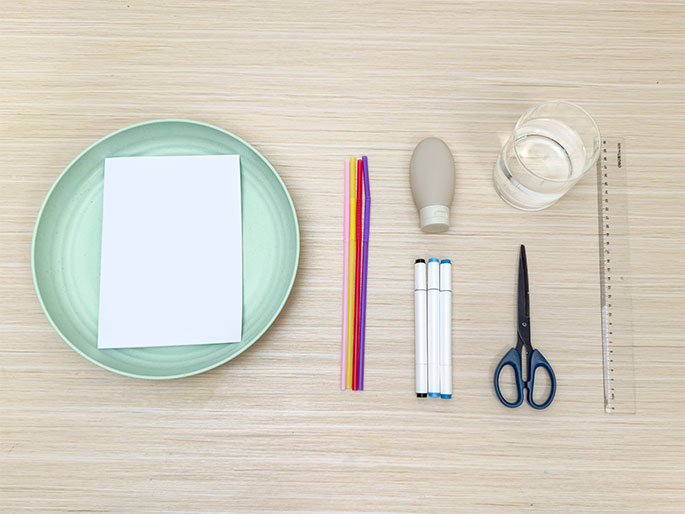 A flat lay arrangement of craft supplies on a light woodgrain surface. The items include a light green plate with a blank white piece of paper on it, a bundle of colorful straws, a small beige bottle or tube, a clear glass of water, a pair of scissors, two white markers with blue and black tips, and a clear plastic ruler. The scene suggests the materials needed for a children's art or science project.