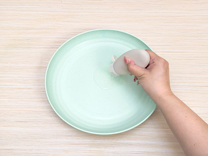 A hand pours a clear liquid from a small, light beige, oval-shaped bottle onto a light green plate. The background is a light woodgrain surface. This suggests a step in a process, likely an art project or experiment, where the liquid is being prepared or measured before being mixed with other materials.