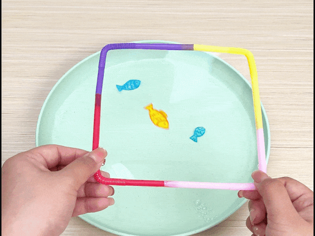 Two hands hold a square frame constructed from connected, colorful plastic straws. The frame floats on the surface of water in a pale green dish, which rests on a light woodgrain table. Inside the floating straw square are three small, colorful fish shapes. The straws are purple, pink, yellow, and pale pink/white. The fish are light blue, yellow/orange, and a smaller light blue. This image likely depicts a playful activity or a child's craft project.