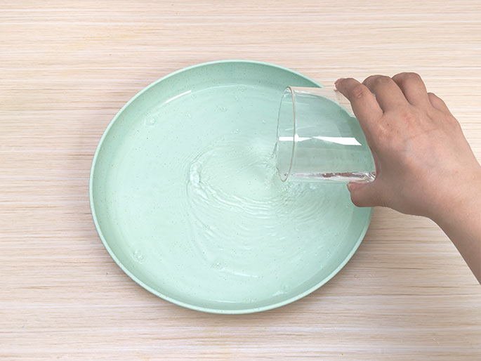A hand pours water from a clear glass into a light green plate. The water is creating ripples in the plate. The background is a light woodgrain surface. This image likely depicts a step in a process, such as preparing for an art project or a science experiment.