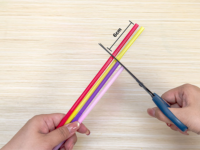 Two hands are shown cutting colorful plastic straws on a light woodgrain surface. One hand holds a group of red, yellow, and light purple straws, while the other hand uses scissors to cut them. A black line indicates a measurement of 6cm, showing the length at which the straws are being cut. This image likely demonstrates a step in a craft project.