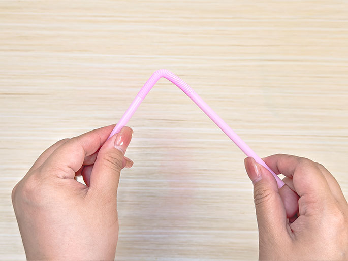 Two hands hold a light pink bendy straw bent into an inverted V shape against a light woodgrain background. The bendable section of the straw is clearly visible at the peak of the V. This image likely demonstrates a step in a craft project where the straw is being prepared or shaped.