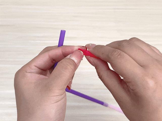 Two hands manipulate colorful plastic straws against a light woodgrain background. One hand holds a purple straw, while the other hand inserts a small, red straw segment into the end of the purple one. This close-up image likely illustrates a step in a craft project involving connecting or joining the straws.