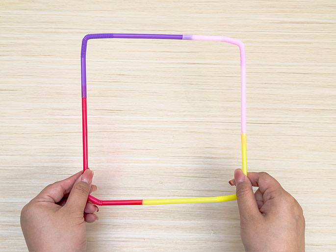 Two hands hold a square frame made of connected, colorful plastic straws against a light woodgrain background. The straws are purple, pink, red, and yellow, creating a vibrant and playful look. This image likely demonstrates the result of a craft project.
