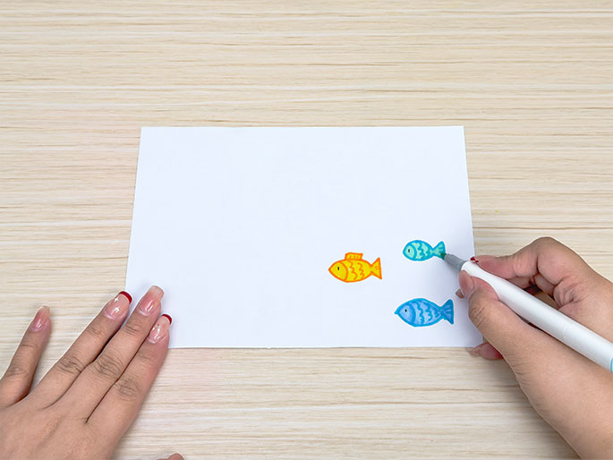 On a piece of white paper placed on a light woodgrain table, a hand draws simple fish illustrations with a marker. There's a small yellow/orange fish, a smaller teal fish, and a larger blue fish being drawn. Another hand lightly holds the paper. The scene suggests a child or someone creating simple artwork.