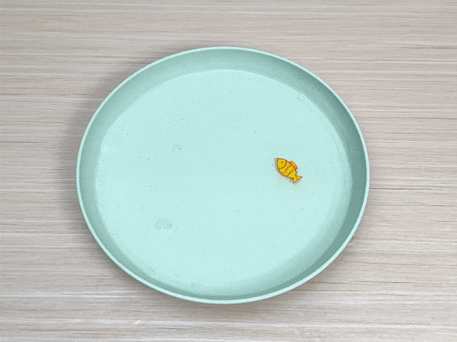 A small, yellow fish-shaped object floats in a shallow, light green dish filled with water. The dish rests on a light woodgrain surface. A few small bubbles are visible in the water. The image is simple and evokes a playful or childlike scene.