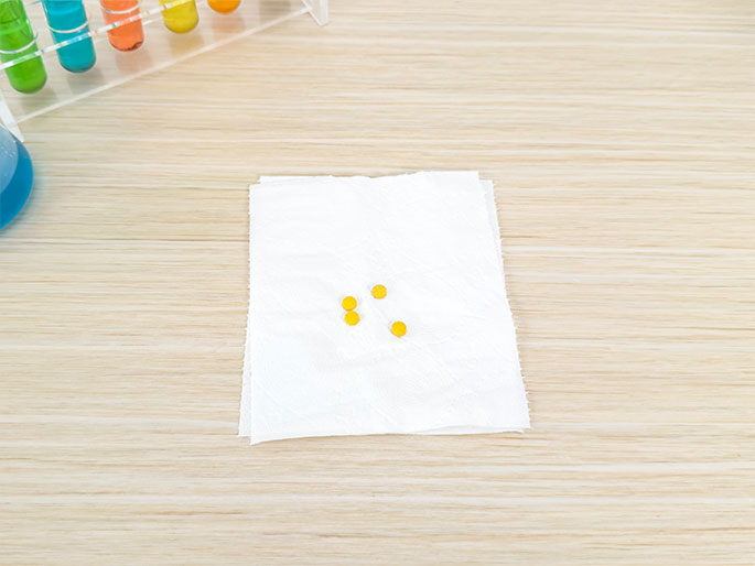 On a light wood-grain table, a folded white paper towel holds a few small, yellow seeds. In the background, a rack of test tubes filled with colorful liquids and a partially visible beaker with blue liquid are visible, suggesting a science experiment or educational setting related to seed germination.