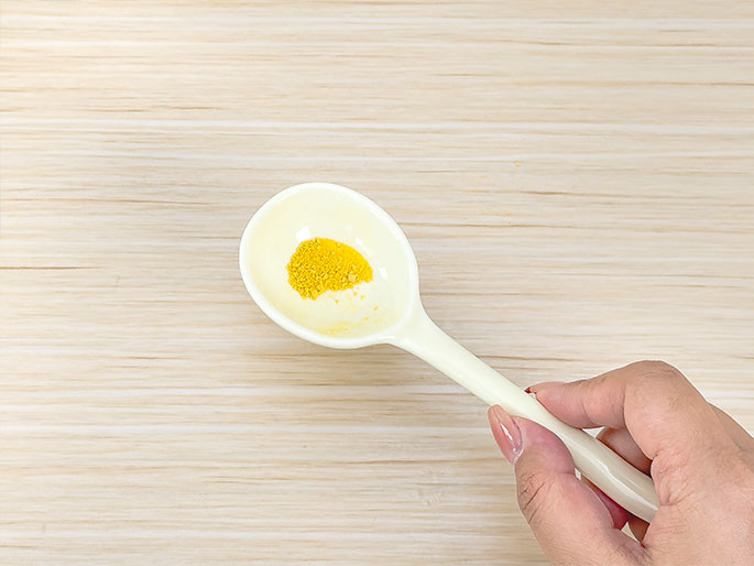 A hand holds a white plastic spoon containing a small amount of yellow powder or spice. The spoon is positioned above a light wood-grain surface. The image suggests cooking, food preparation, or possibly a scientific experiment involving a powdered substance.