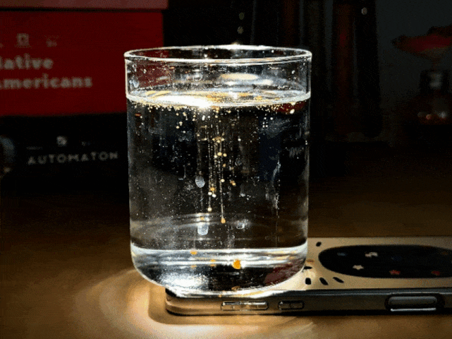 A clear glass of water sits on top of a cell phone. Small, gold-colored particles drift down through the water, settling at the bottom of the glass. The light source appears to be from below, illuminating the glass and creating a bright circle on the surface beneath it. The background is dark and out of focus, with the words Native Americans and Automaton partially visible on what appears to be a book or box.