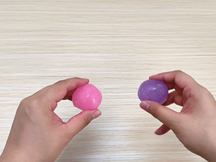 Two hands hold a small, bright pink ball and a sparkly purple ball. Both balls appear to be made of a soft, rubbery, or gel-like material. The hands release the balls, and they bounce on a light wooden surface. The video showcases the bouncy and tactile qualities of two colorful balls.