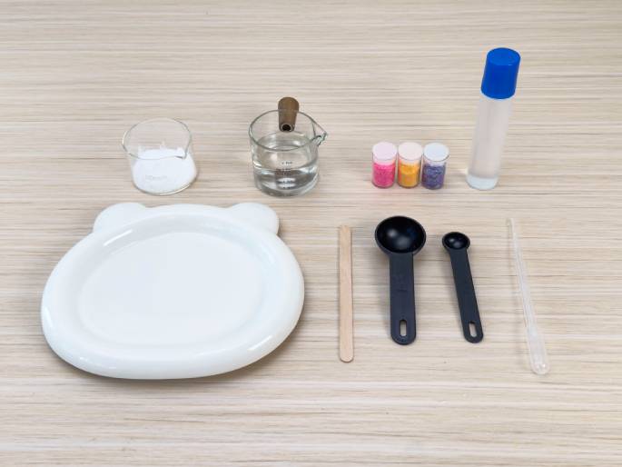 An assortment of materials, likely for a science experiment or craft project, is arranged on a light woodgrain table. From left to right, the items include: a small glass beaker with a white powder, a glass measuring cup with a wooden handle containing clear liquid, three small containers of colorful glitter or beads (pink, orange, and purple), a clear bottle with a blue cap containing a clear liquid (possibly glue), a white plate with bear-ear-shaped protrusions, a wooden stirring stick, two black measuring spoons of different sizes, and a clear pipette or dropper. This image likely depicts the ingredients and tools gathered before starting the activity.