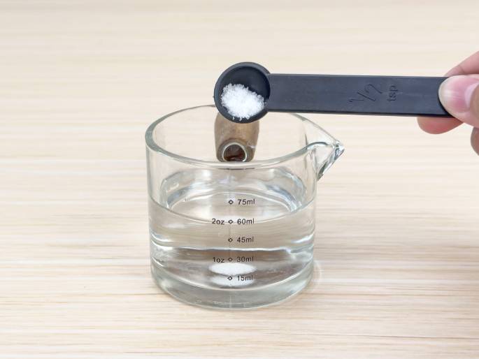 A hand holds a black 1/2 teaspoon measuring spoon filled with white granules, likely salt or sugar, over a clear glass measuring cup containing water. The cup sits on a light woodgrain table, and the water level is between the 15ml and 30ml markings. A small, brown cylindrical object, possibly a cork or wooden dowel, floats in the water. The image likely depicts a step in a science experiment demonstrating density or solubility.