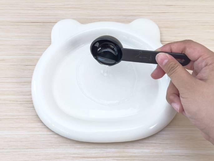 A hand pours a clear liquid from a small, black measuring spoon onto a white plate with bear-ear-shaped protrusions. The plate sits on a light woodgrain table, and a small puddle of the liquid is forming on the plate's surface. This image likely depicts a stage in a recipe or experiment where a liquid ingredient is being added.