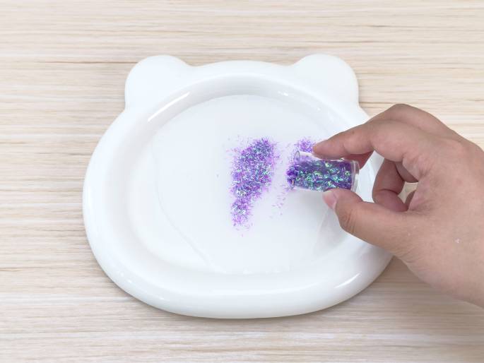 A hand pours iridescent purple glitter from a small, clear container onto a white plate with bear-ear-shaped protrusions. Some glitter has already been poured onto the plate, and the plate sits on a light woodgrain table. This image likely depicts adding glitter to a craft project, perhaps slime making or another similar activity.