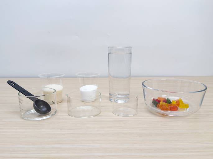 An assortment of ingredients and equipment are arranged on a light woodgrain table against a plain off-white background. From left to right, there's a small clear measuring cup with a black measuring spoon inside, two slightly larger clear containers holding a granulated substance (likely sugar and salt), a tall glass of water, two small empty clear containers, and a clear glass bowl containing a handful of colorful gummy bear candies. This setup suggests preparation for a science experiment or kitchen activity, possibly involving dissolving the gummies or creating a solution.