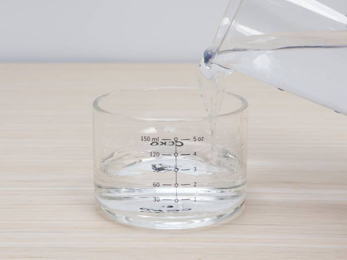 Water is being poured from a clear glass beaker into a clear glass measuring cup on a light woodgrain table. The measuring cup has milliliter and ounce markings, and the water level is rising as it's poured. The background is a plain off-white wall. This image likely depicts a step in a recipe or experiment where a precise amount of water is required.