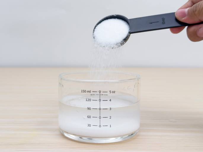A hand holds a black measuring spoon filled with white granulated material, likely sugar or salt, pouring it into a clear glass measuring cup containing water. The measuring cup sits on a light woodgrain table and has milliliter and ounce markings. The background is a plain off-white wall. This image likely depicts a step in a recipe or experiment requiring dissolving a specific amount of a dry ingredient in water.