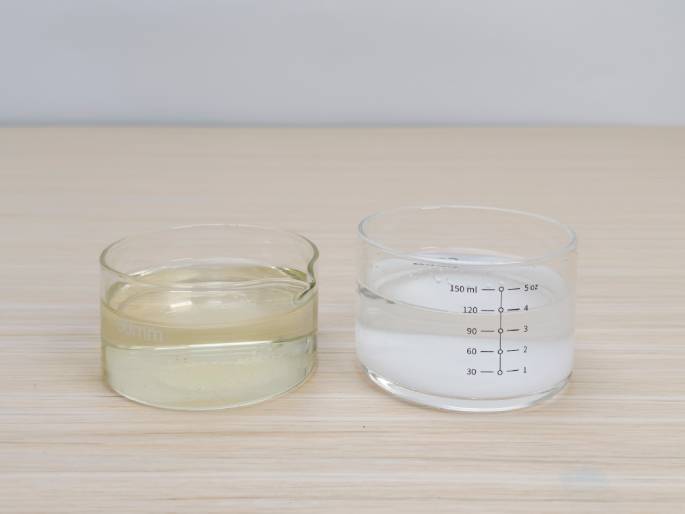 Two clear glass containers sit on a light woodgrain table against a plain off-white background. The container on the left is a short, wide beaker holding a yellowish liquid, possibly oil or another similar substance. The container on the right is a measuring cup with milliliter and ounce markings, containing a clear liquid, likely water or a water-based solution. This image likely depicts the ingredients for a science experiment or a recipe involving the combination of these two liquids.