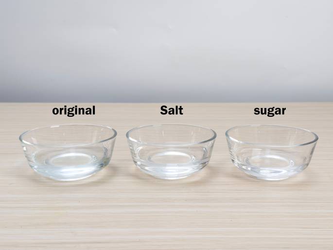 Three small, clear glass bowls containing clear liquids are arranged on a light woodgrain table against a plain off-white background. The bowls are labeled from left to right: original, Salt, and Sugar. This setup suggests an experiment or comparison of how different solutes (salt and sugar) affect a liquid, likely water. The original bowl presumably contains the liquid without any added solutes.