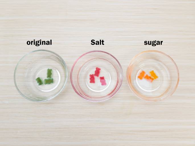 Three clear glass bowls are arranged on a light woodgrain table against a plain off-white background. Each bowl contains a few gummy bear candies. From left to right, the bowls are labeled original, Salt, and Sugar. The gummy bears in the original bowl are green. The Salt bowl contains red gummy bears, and the Sugar bowl has orange gummy bears. This arrangement suggests an experiment to observe the effects of salt and sugar solutions on gummy bears.