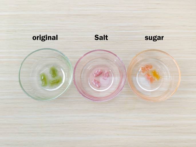 Three clear glass bowls sit on a light woodgrain table against a plain off-white background. Each bowl contains a few gummy bears, and they are labeled from left to right as original, Salt, and Sugar. The gummy bears in the original bowl appear slightly swollen and maintain their original green color. The gummy bears in the Salt bowl are noticeably larger and a lighter, almost translucent pink color. The gummy bears in the Sugar bowl are also enlarged, but less so than those in the salt bowl, and have a pale orange hue. This image likely illustrates the results of an experiment demonstrating the effects of different solutions on gummy bears.