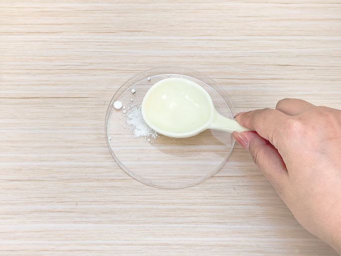 A hand holds a small, white plastic measuring spoon over a clear dish on a light wood-grain table. A small pile of white powder and a few fragments of a crushed white tablet are visible in the dish. The scene suggests the preparation of an experiment, possibly involving mixing or measuring ingredients.