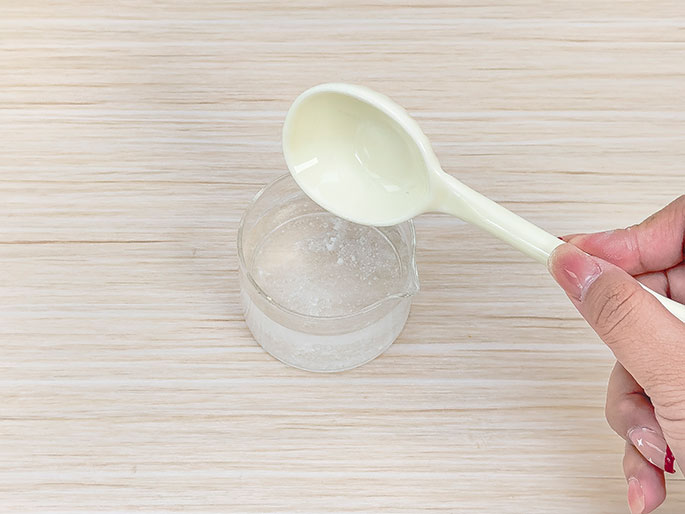 A hand holds a white plastic spoon above a clear glass beaker containing a clear liquid and some white, dissolved particles or powder. The setup, placed on a light wood-grain surface, suggests a science experiment or activity involving dissolving a substance in liquid.