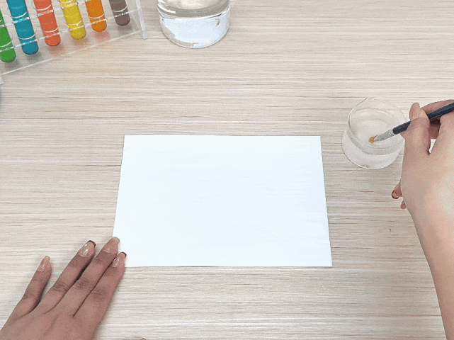 Two hands are positioned near a blank white sheet of paper on a light wooden table. One hand holds the paper while the other dips a small paintbrush into a clear container filled with a light-colored liquid. A rack of colorful test tubes and a clear glass of water are also visible in the background. The arrangement suggests an art project or a science experiment involving painting or applying a liquid to the paper.