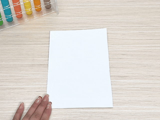 A hand is positioned to hold or adjust a blank white piece of paper on a light wood-grain table. In the top left corner, a rack of colorful test tubes is visible, suggesting a setting related to a science experiment, art activity, or educational demonstration.