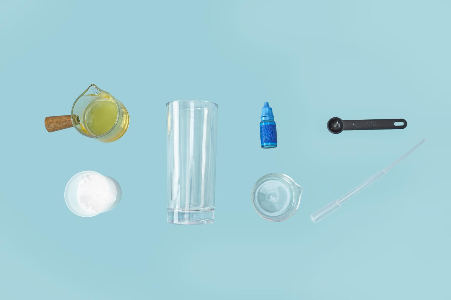 Against a pale blue background, various items are neatly arranged for a science experiment, likely to create a homemade lava lamp. These include a small glass jug containing a yellowish liquid (likely oil), a tall clear glass, a small bottle of blue liquid (likely food colouring), a small measuring spoon, a small empty glass bowl, and a pipette. A small container of white powder (possibly salt or baking soda) is also present. The items are spaced evenly, creating a visually appealing and organised presentation, ready for the experiment to begin.