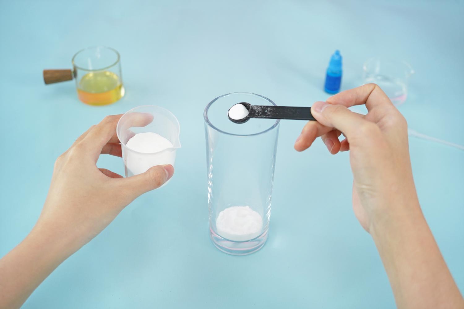 A person's hands are shown adding a white powder, likely baking soda, to a clear glass using a small black measuring spoon. A small plastic measuring cup containing more of the white powder is held in the other hand. In the background, a small glass jug of yellowish liquid (oil) and a small bottle of blue liquid (food colouring) are visible, along with a small empty beaker. The scene is set against a light blue surface. The image depicts a step in the process of making a homemade lava lamp, focusing on the addition of a key ingredient.