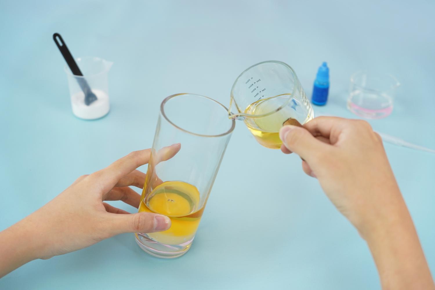 Two hands carefully pour a yellowish liquid, likely oil, from a small measuring jug into a tall, clear glass. A layer of white substance is already visible at the bottom of the glass. Other items are visible in the background, including a small container of white powder (possibly baking soda), a small bottle of blue liquid (food colouring), and a small black spatula. The scene is set against a light blue surface. The image depicts a step in the process of making a homemade lava lamp, showing the addition of oil to the base.