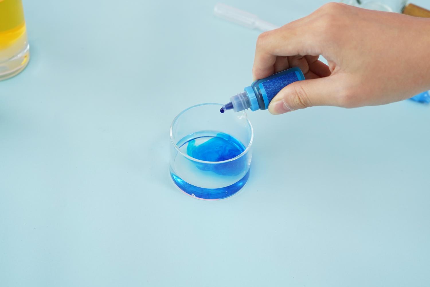 A hand is carefully adding blue food colouring from a small bottle into a clear glass bowl. A small amount of blue liquid is already present in the bowl. The background is a plain, light blue surface. The image shows a step in the process of preparing ingredients for a homemade lava lamp, focusing on the addition of colour to one of the liquids. The overall style is clean and instructional.