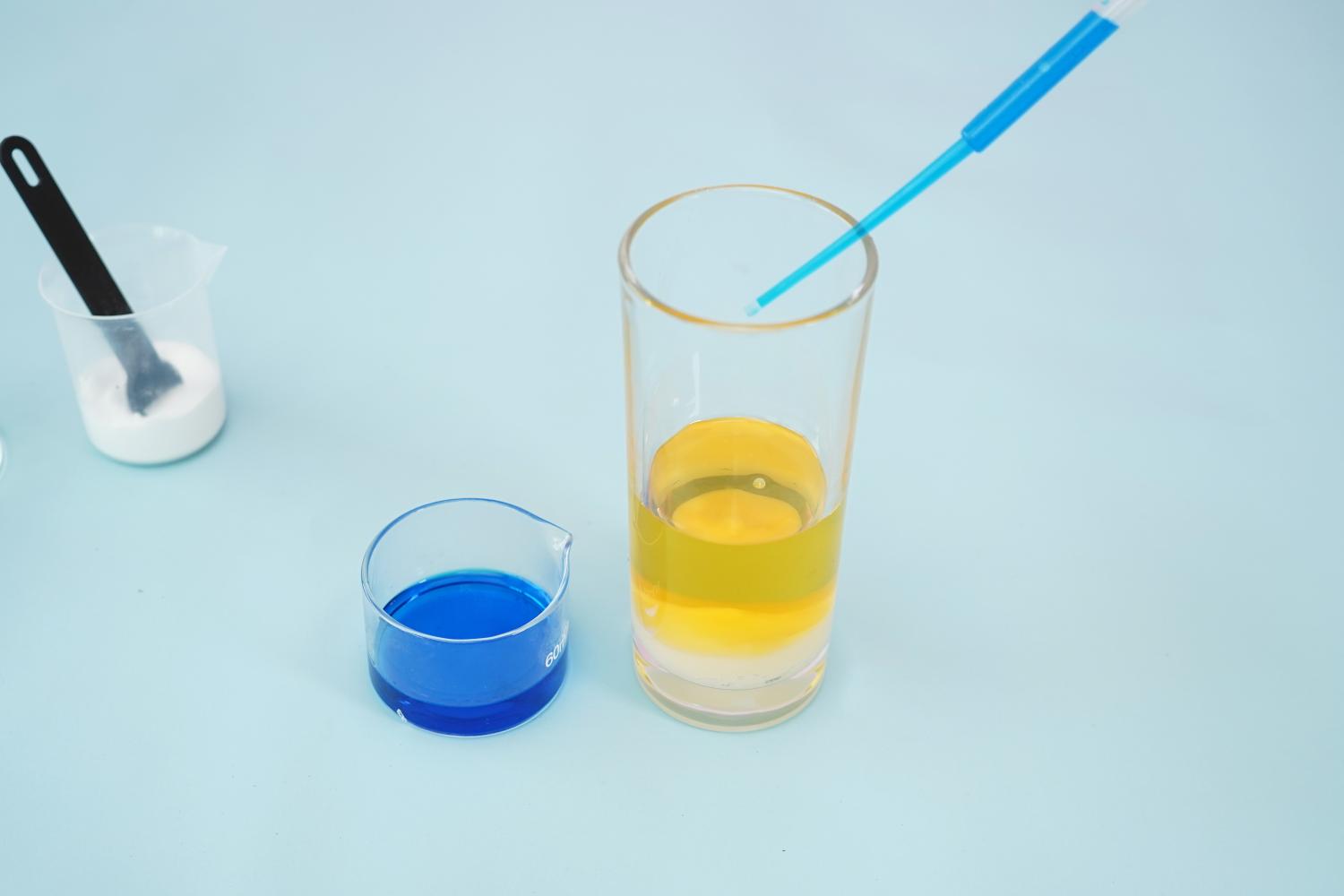 A tall, clear glass contains a layer of yellowish liquid (oil) on top of a layer of white (water and baking soda). A blue liquid (coloured water) is in a smaller beaker to the left. A pipette, containing a small amount of blue liquid, rests in the tall glass. A small measuring beaker containing white powder (baking soda) and a small black spatula are visible in the background. The entire setup rests on a pale blue surface. The image shows a step in the process of creating a homemade lava lamp, illustrating the layering of liquids and the addition of coloured water using a pipette.