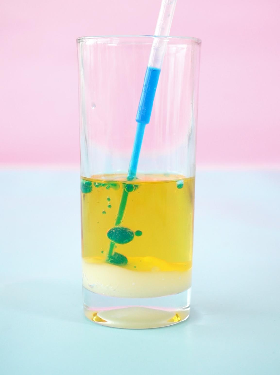 A tall, clear glass, standing on a pale blue surface against a pale pink background, contains a homemade lava lamp. The glass shows distinct layers: a base of white liquid, topped by a layer of yellow oil, within which are several small, green droplets. A blue pipette is inserted into the mixture, suggesting the process of adding the coloured liquid. The image focuses on the layered effect and the process of creating the lava lamp, highlighting the contrast between the colours and the immiscibility of the liquids.
