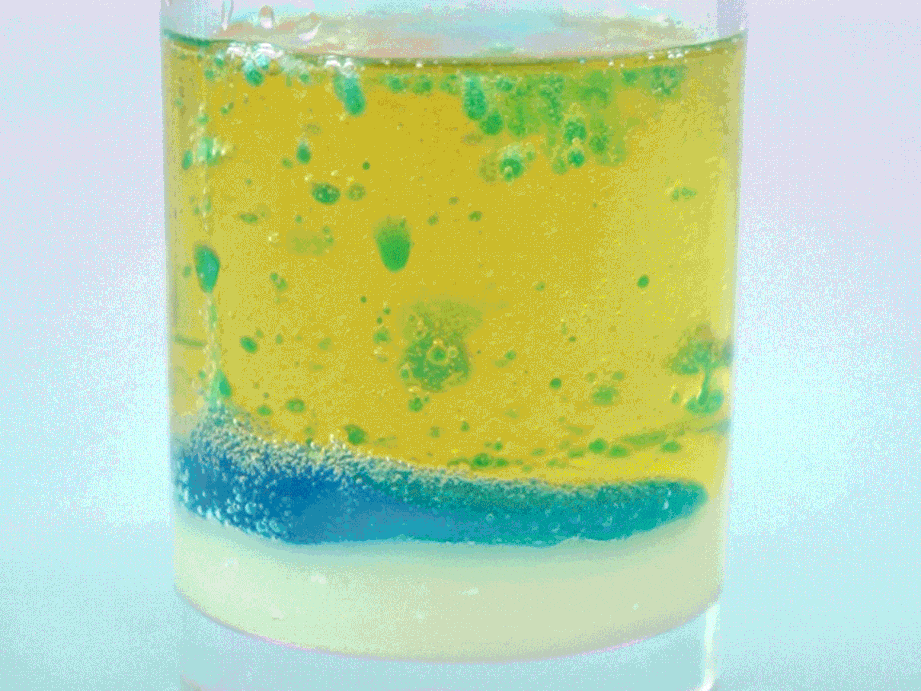 A clear glass, possibly a small tumbler or beaker, contains three distinct layers of liquid. A pale, almost translucent layer forms the base, topped by a thin band of vibrant blue. The largest layer, a rich yellow, sits on top and contains small droplets of green, creating a visually interesting effect. This layered arrangement suggests a density experiment, where liquids of varying densities separate and settle according to their weight.