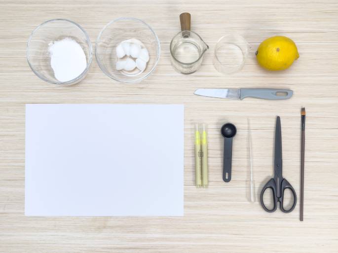 Various materials are laid out on a light woodgrain table, seemingly for a science experiment or art project. Two clear glass bowls contain white substances: one appears to hold baking soda or a similar powder, while the other contains several cotton balls. A small glass beaker with a wooden handle holds a clear liquid. An empty clear glass jar, a whole lemon, a small knife, two yellow markers or paintbrushes with caps, a small black measuring spoon, a clear pipette or dropper, a pair of scissors, a paintbrush, and a blank white sheet of paper complete the arrangement. This flat lay suggests preparation for an activity involving invisible ink, citrus-based reactions, or a similar scientific or artistic endeavor.