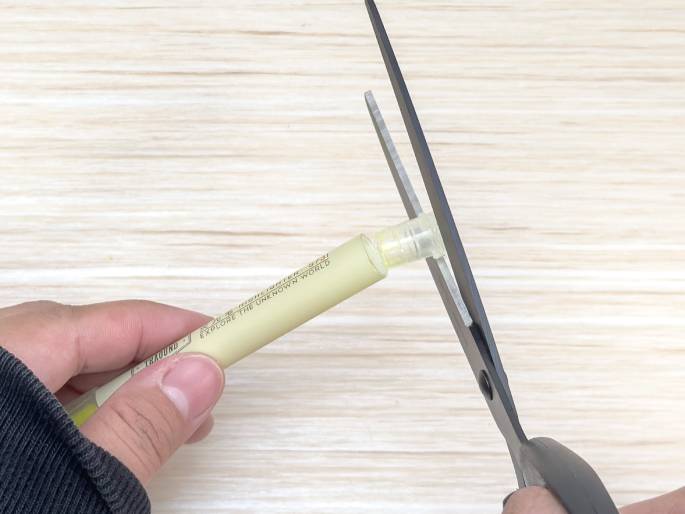 A hand holds a pale yellow marker horizontally above a light woodgrain surface. The marker is being cut near its tip with a pair of scissors held in another hand. The focus is on the action of cutting the marker, with the scissors blades positioned to sever the plastic casing. The marker has some text visible, suggesting it's an art or craft supply. This image likely demonstrates a step in a DIY project or experiment involving modifying a marker, perhaps to extract ink or create a custom nib.