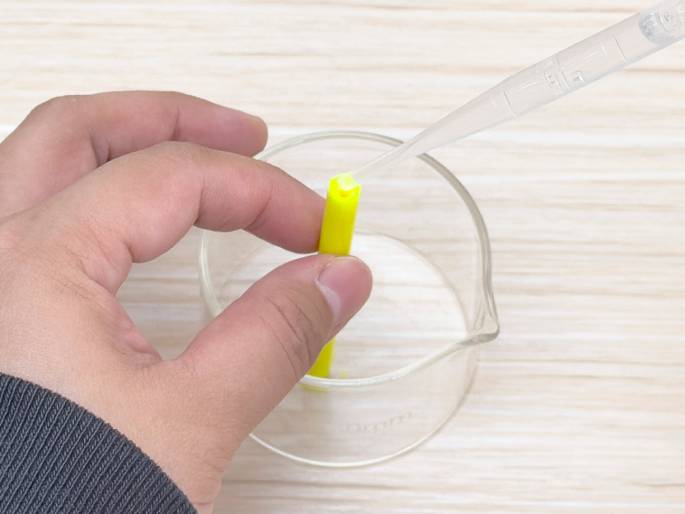 A hand holds a bright yellow marker upright over a clear glass beaker on a light woodgrain table. A plastic pipette dispenses drops of a clear liquid onto the exposed, cut end of the marker. The beaker appears empty or contains a small amount of a clear solution. This close-up image suggests a scientific experiment or DIY project involving the absorption of liquid by the marker's inner core, possibly to create a custom color or effect.