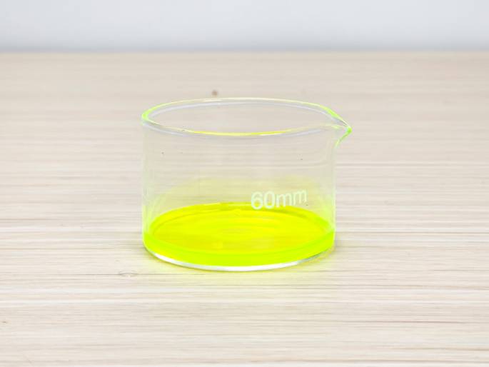 A small, clear glass beaker sits on a light woodgrain table against a plain off-white background. The beaker contains a vibrant, fluorescent yellow liquid, filling the bottom portion of the vessel. A 60mm marking is visible on the beaker's side, likely indicating its diameter. This simple image suggests a scientific experiment or demonstration involving a chemical reaction or solution, highlighting the vivid color of the liquid.