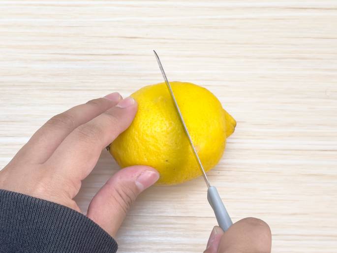 Two hands are shown in the process of cutting a lemon on a light woodgrain surface. One hand holds the lemon steady while the other uses a small knife with a gray handle to slice through it. The lemon is bright yellow and appears ripe. The focus is on the act of cutting, with the knife positioned midway through the lemon. This image likely depicts a step in a recipe or experiment requiring fresh lemon juice.